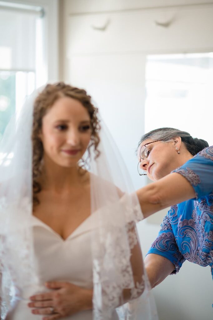 bride in wedding dress and veil smiles while her mother adjusts the dress